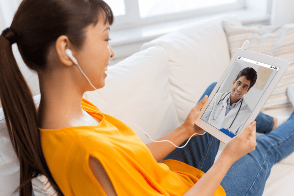 A woman sitting comfortably on a couch, busy with a video with her doctor on her tablet.