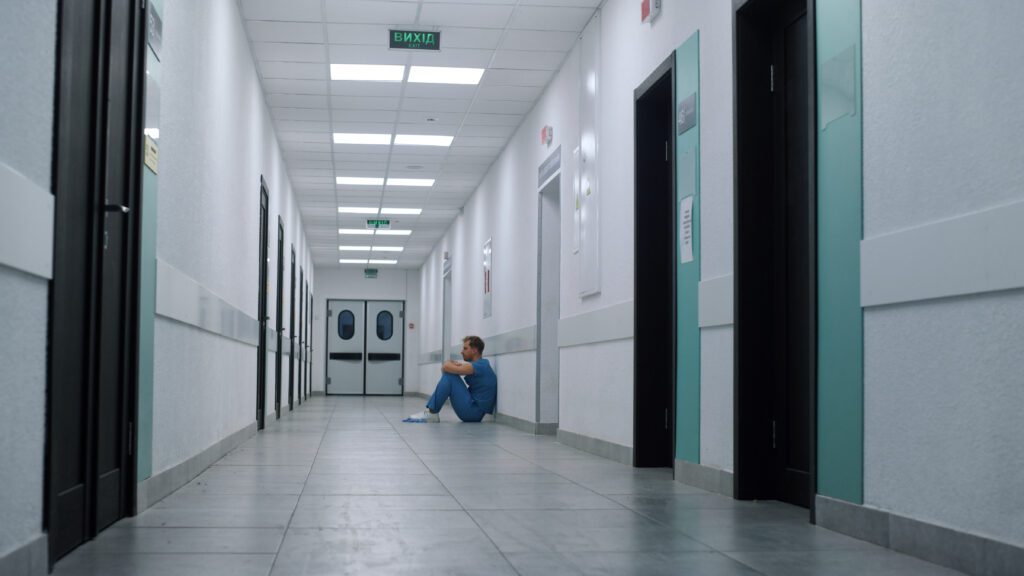 Overwhelmed medicine worker sitting floor long ambulance hallway. Alone man doctor resting empty hospital corridor for transporting critically ill patients. Tired unknown medic relaxing after surgery.