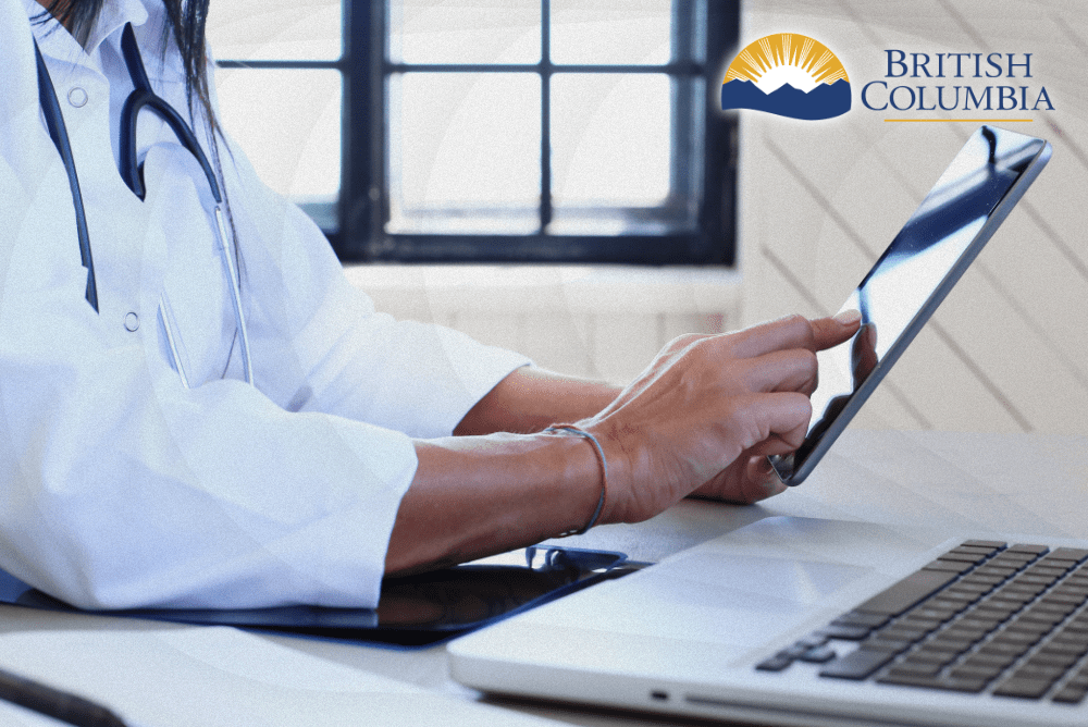 A medical professional in a white coat uses a tablet, with a laptop and notebook on a wooden desk. British Columbia logo displayed.