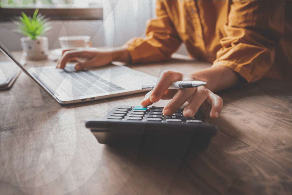 Person using a laptop and calculator on a wooden desk, typing with one hand and holding a pen in the other.