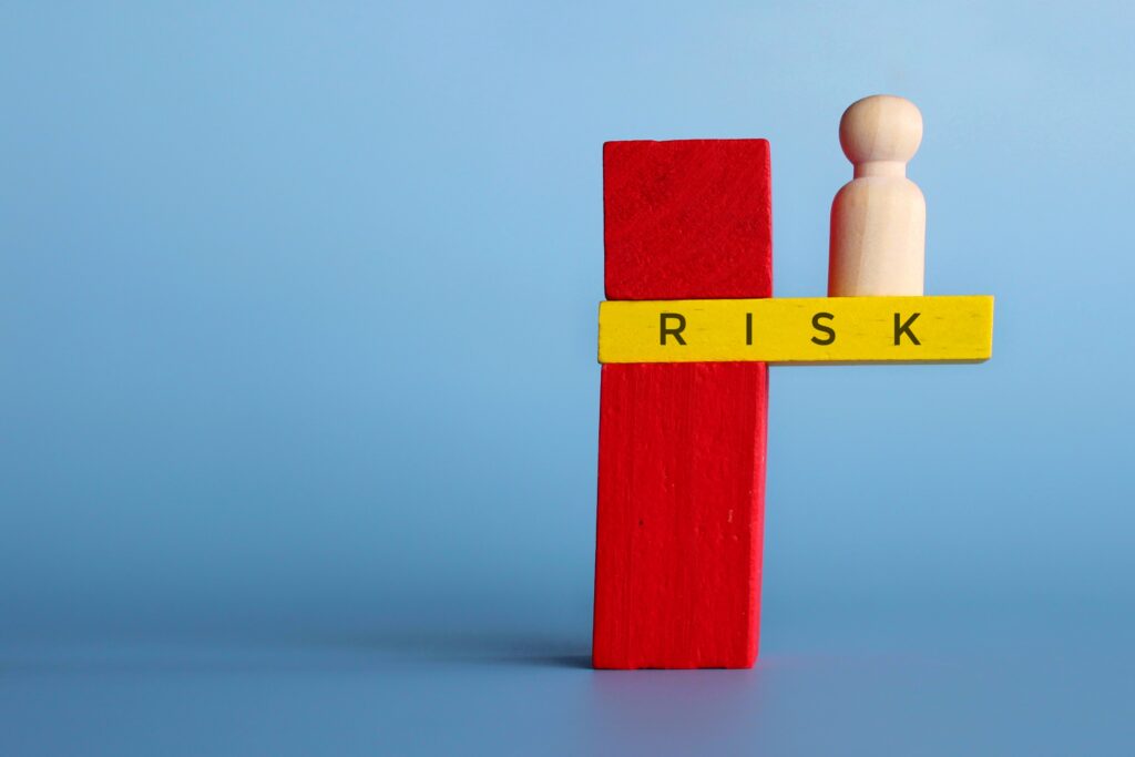 A red block stands upright, supporting a yellow beam labeled "RISK" with a wooden figure on top against a blue background, symbolizing balance and caution.