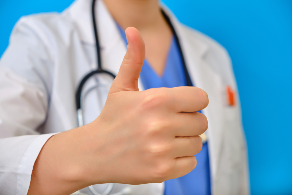 A doctor in a white coat and blue scrubs gives a thumbs-up, conveying approval. A stethoscope hangs around the neck against a blue background.