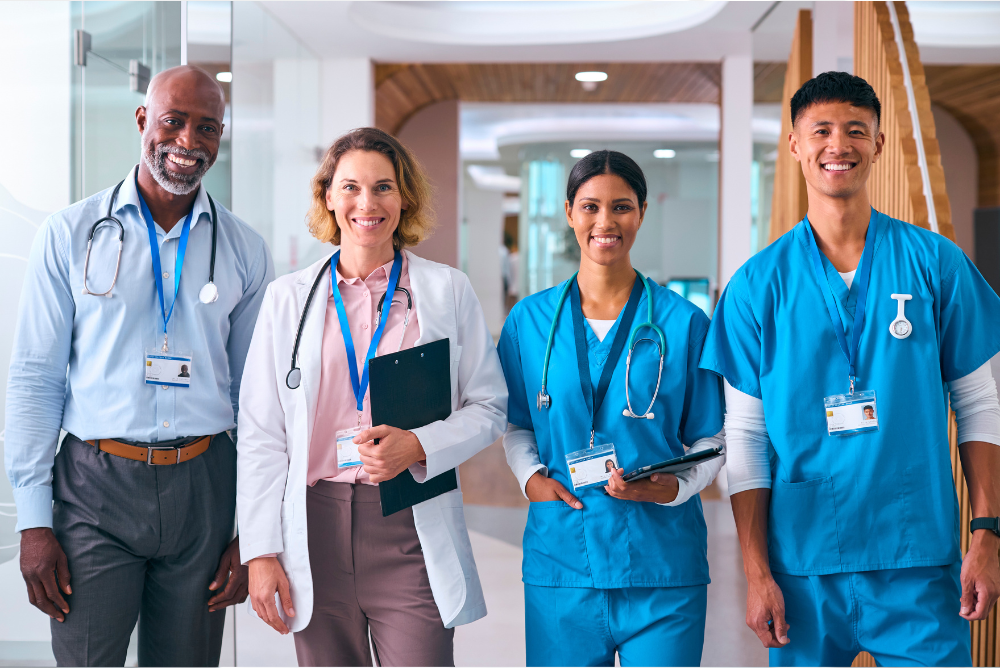 Four healthcare professionals, two in blue scrubs and two in white coats, smiling and standing in a hospital hallway. The mood is professional and welcoming.