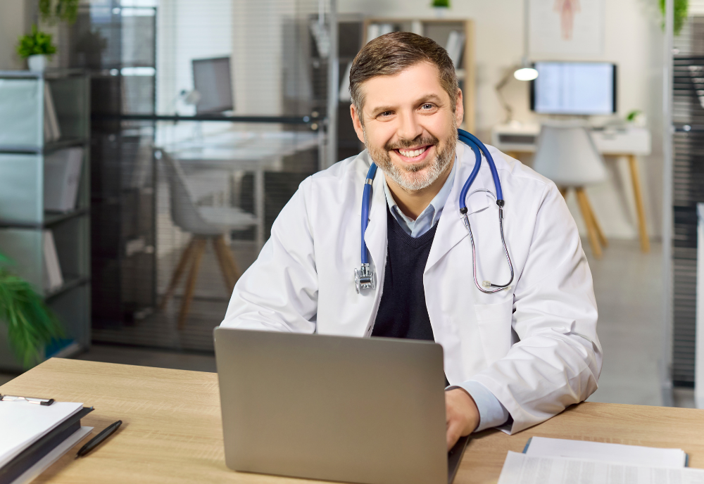 A smiling doctor with a beard, wearing a white coat and stethoscope, sits at a desk with a laptop in a bright, modern office setting.