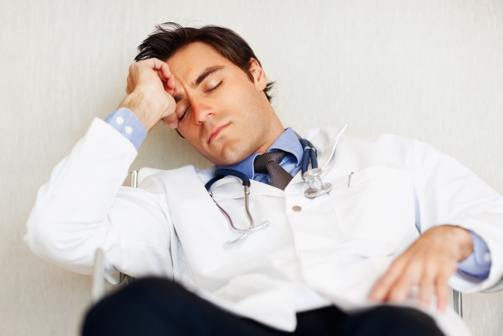 A tired doctor in a white coat with a stethoscope naps in a chair, leaning against the wall. His expression shows exhaustion, reflecting a demanding workday.