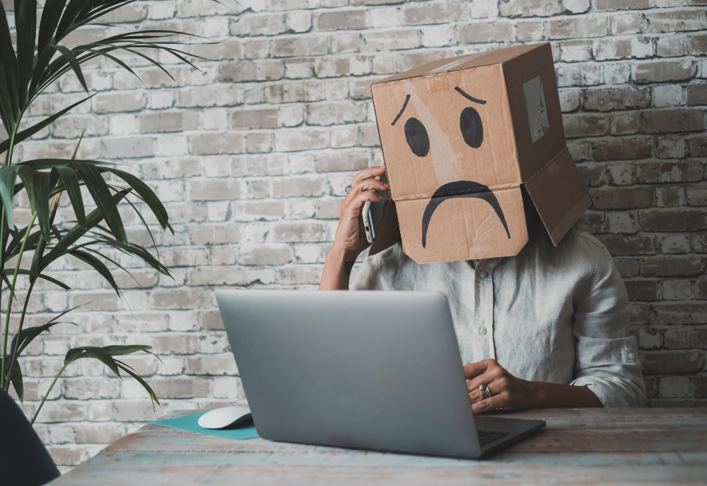 Person with a cardboard box depicting a sad face over their head, talking on the phone, seated at a desk with a laptop. The setting is casual and quirky.