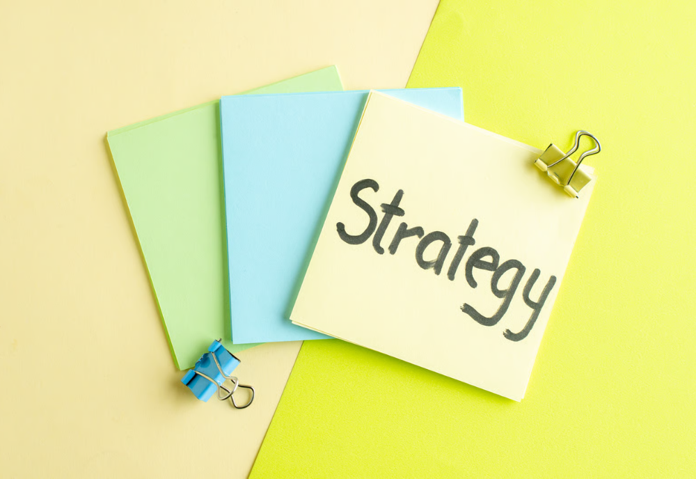 A strategy written on a yellow sticky note, clipped with blue, on a vibrant yellow and green backdrop.