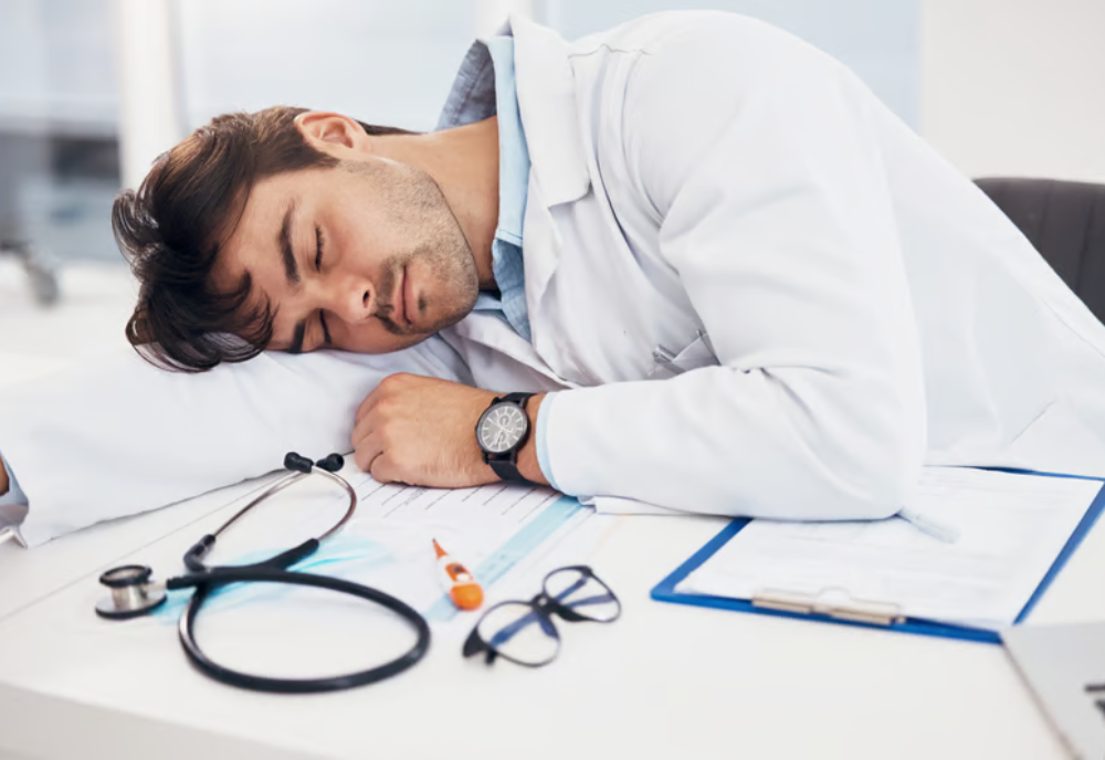 A man in a white coat is asleep at a desk, his head down on the surface, with various documents and a laptop nearby.