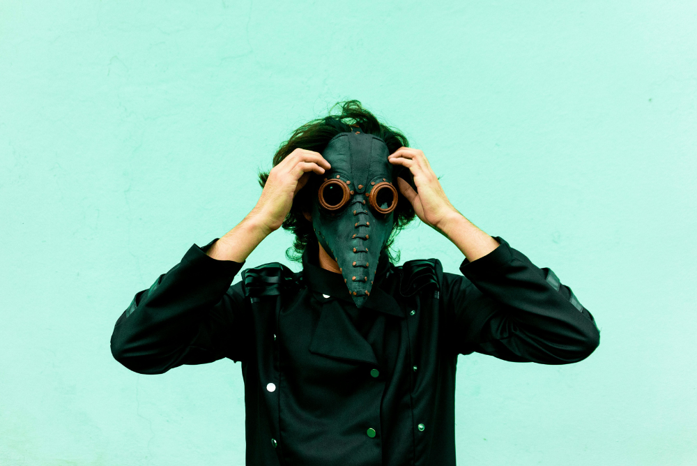 A man with a bird mask on his head, stands in a simple setting.